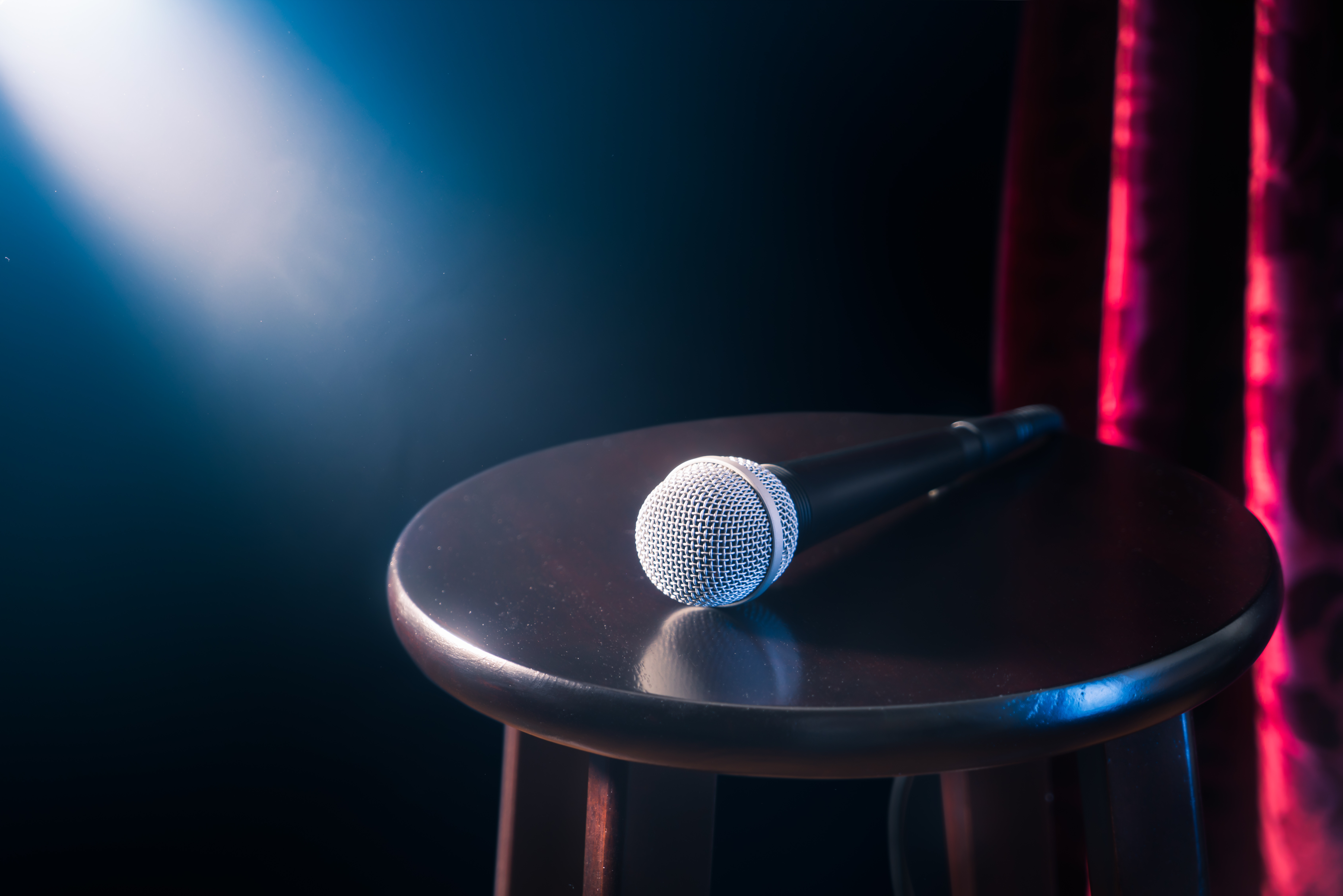 Microphone resting on a wooden stool under stage spotlight, framed by red velvet curtains